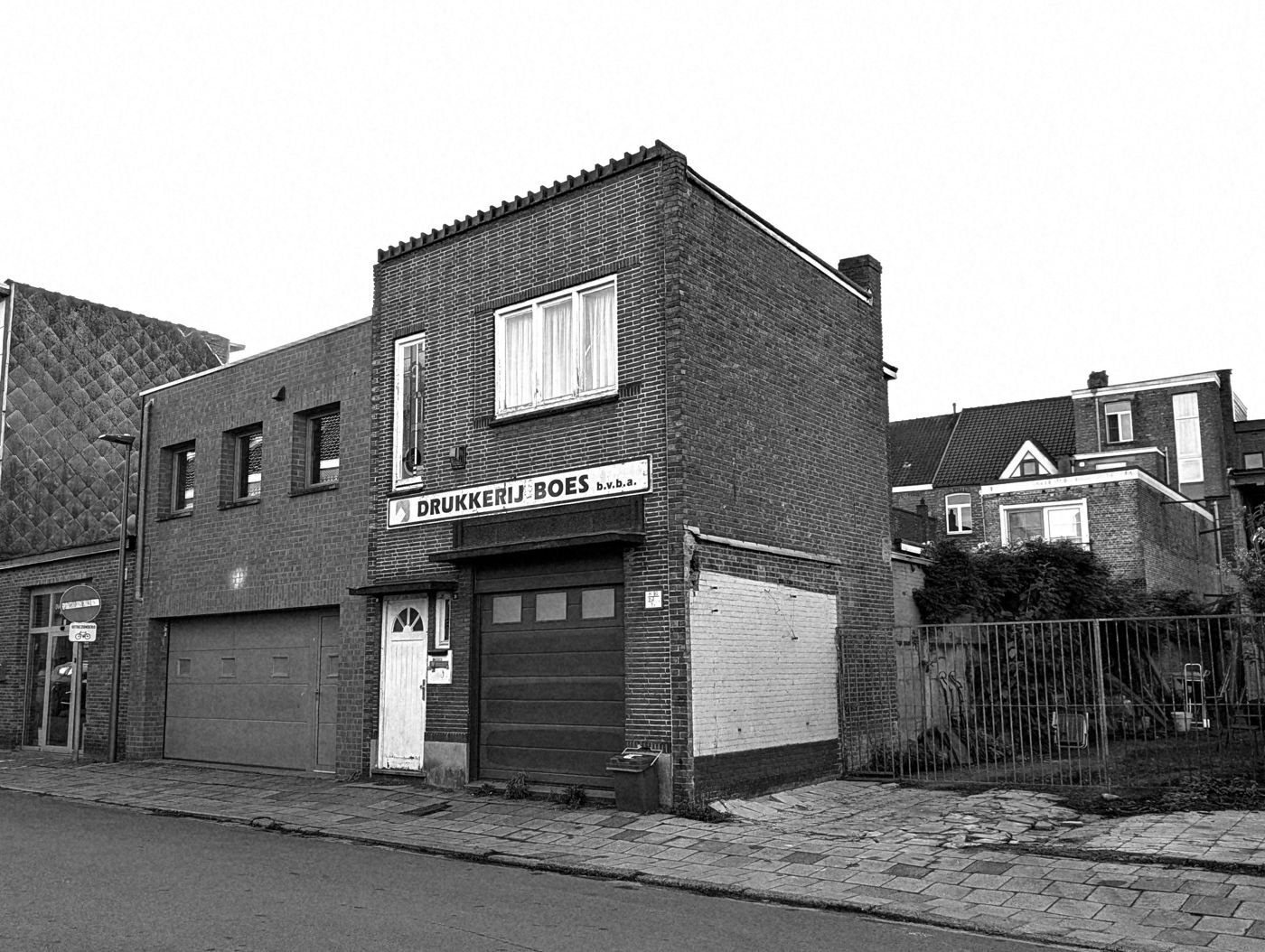 Bakstenen gevel van een oude drukkerij met verbleekt uithangbord, witte voordeur en gesloten garagepoort