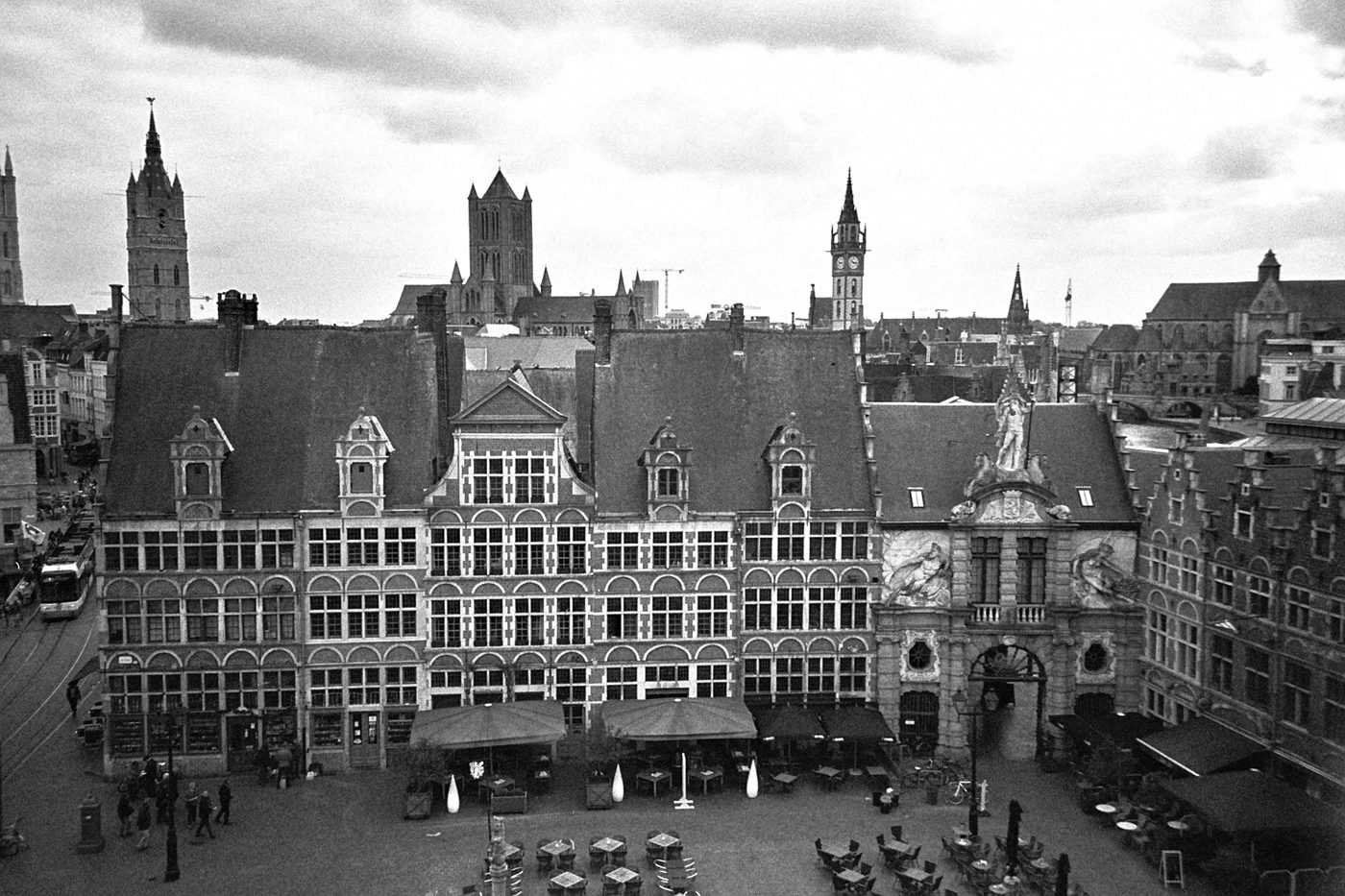 Zicht van bovenaf op een plein in Gent met trapgevels, terrassen en de torens van de binnenstad onder een bewolkte lucht