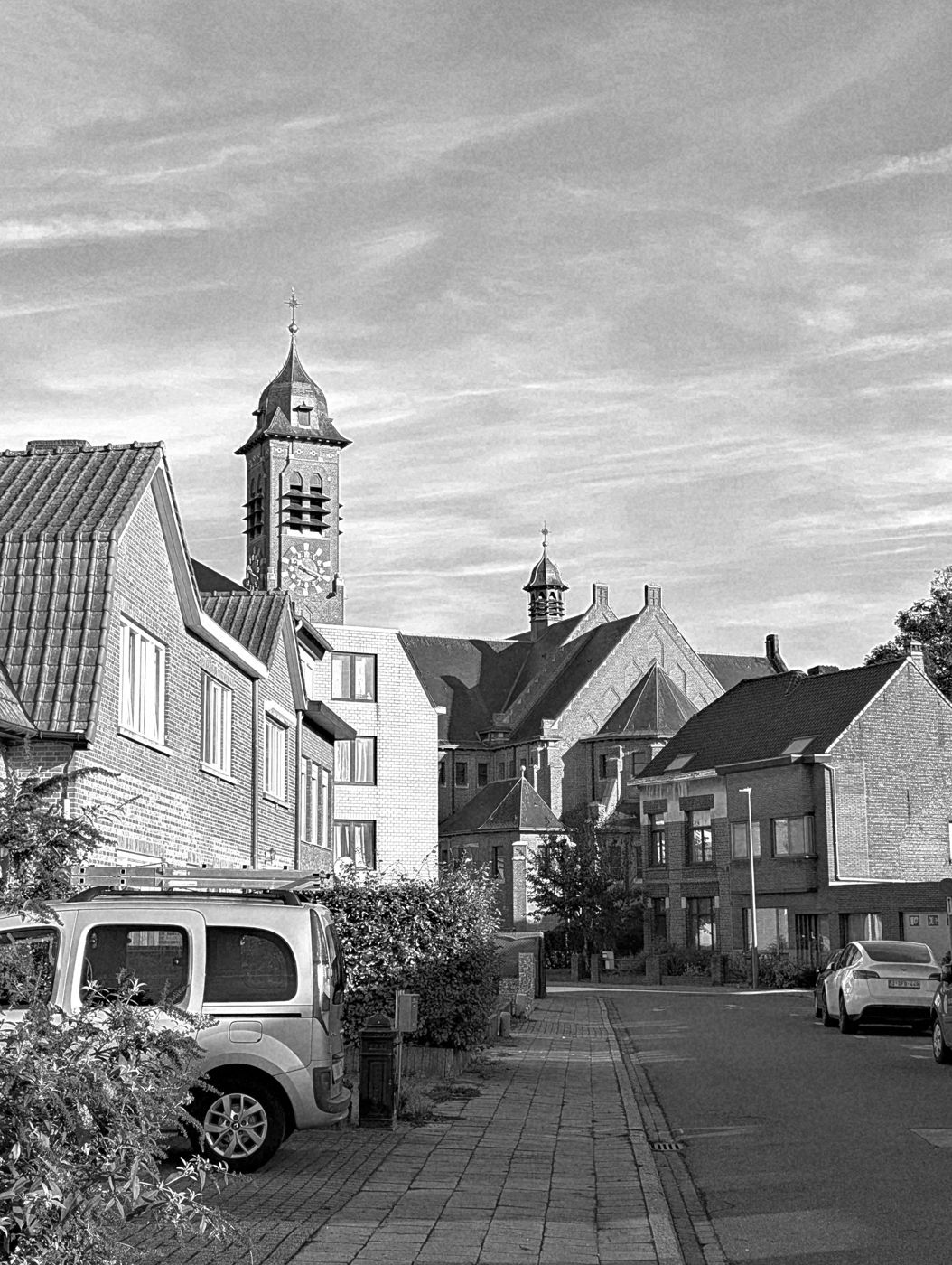 Straat met bakstenen huizen, daarachter een kerk met klokkentoren onder een gestreepte wolkenlucht